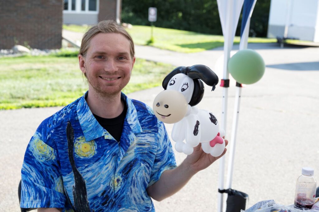 Smiling man in a blue patterned shirt holds a black-and-white cow puppet at an outdoor event.