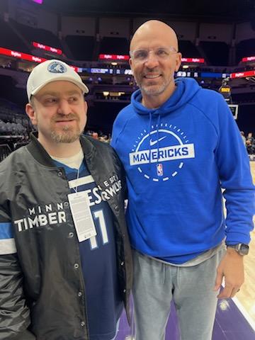 Two men pose together on a basketball court, one wearing a Dallas Mavericks hoodie and the other in a cap and jacket.