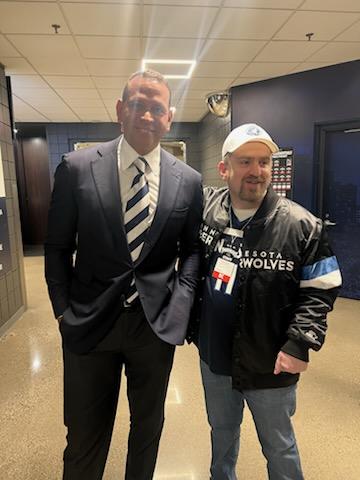 Two men pose for a photo in a hallway, one wearing a dark suit and striped tie, the other in a black jacket and white cap.