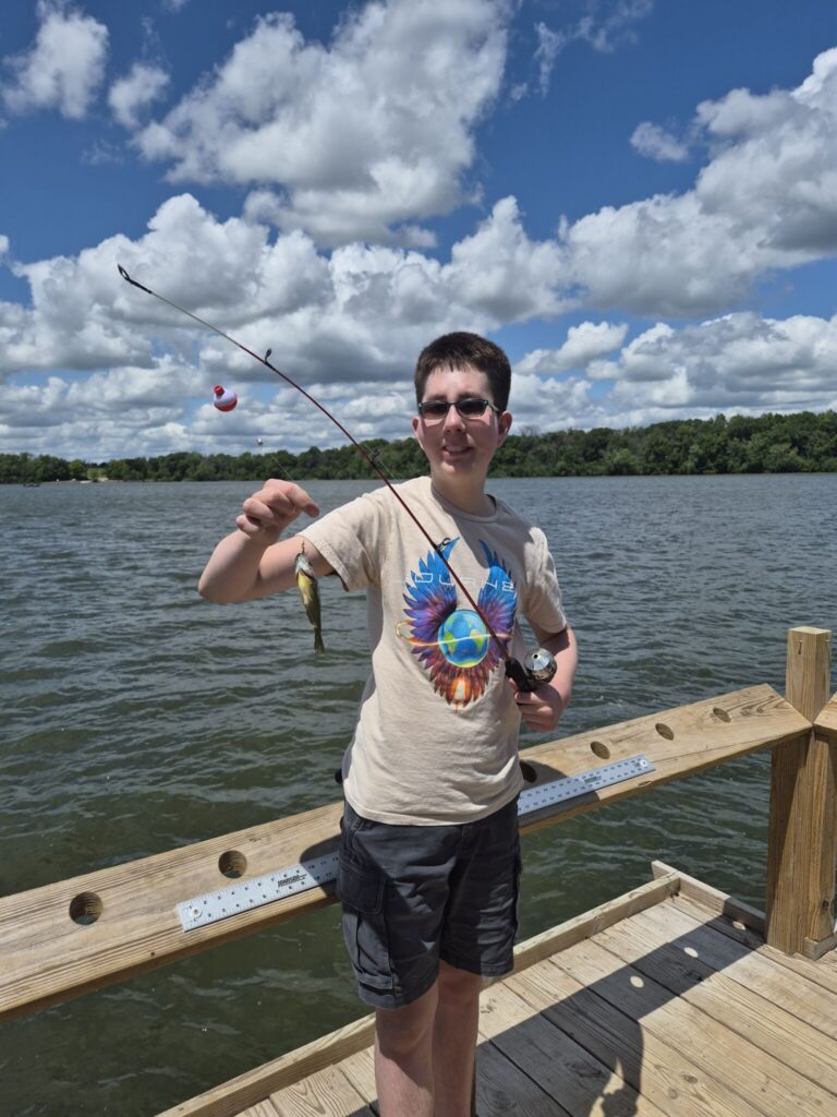 Person standing on a wooden dock beside a lake, holding up a small fish they just caught.