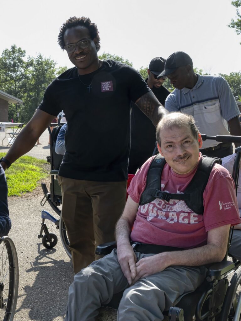 A man in a pink shirt sits in a wheelchair outdoors, with two companions standing beside him on a sunny day.