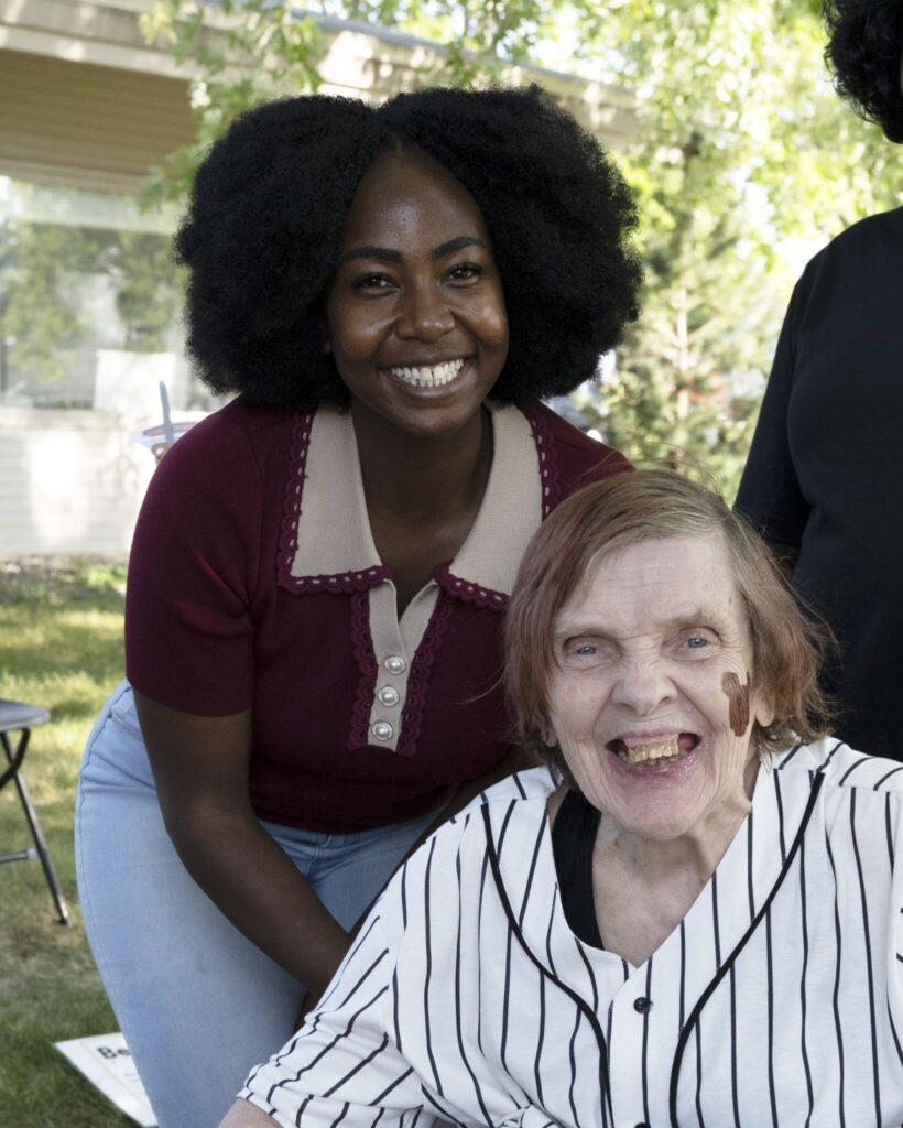 Two women smiling outdoors, one seated in a striped shirt and the other standing behind her.