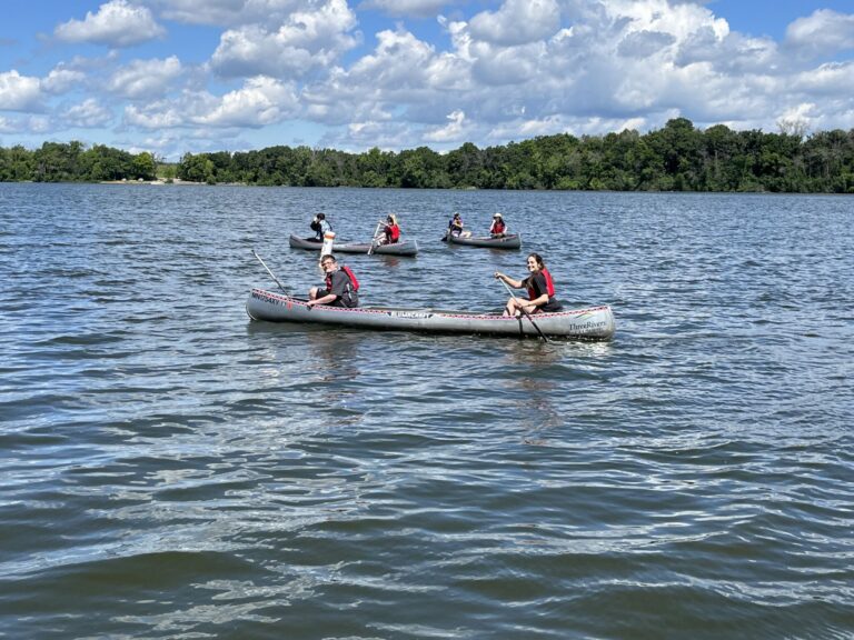 Two small boats with people wearing life jackets paddling on a lake, with a tree-lined shoreline and a blue sky.
