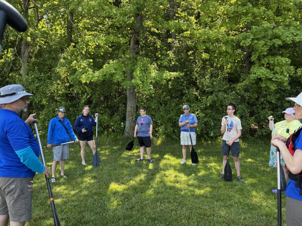A group of people stands in a circle on a grassy area beneath trees.