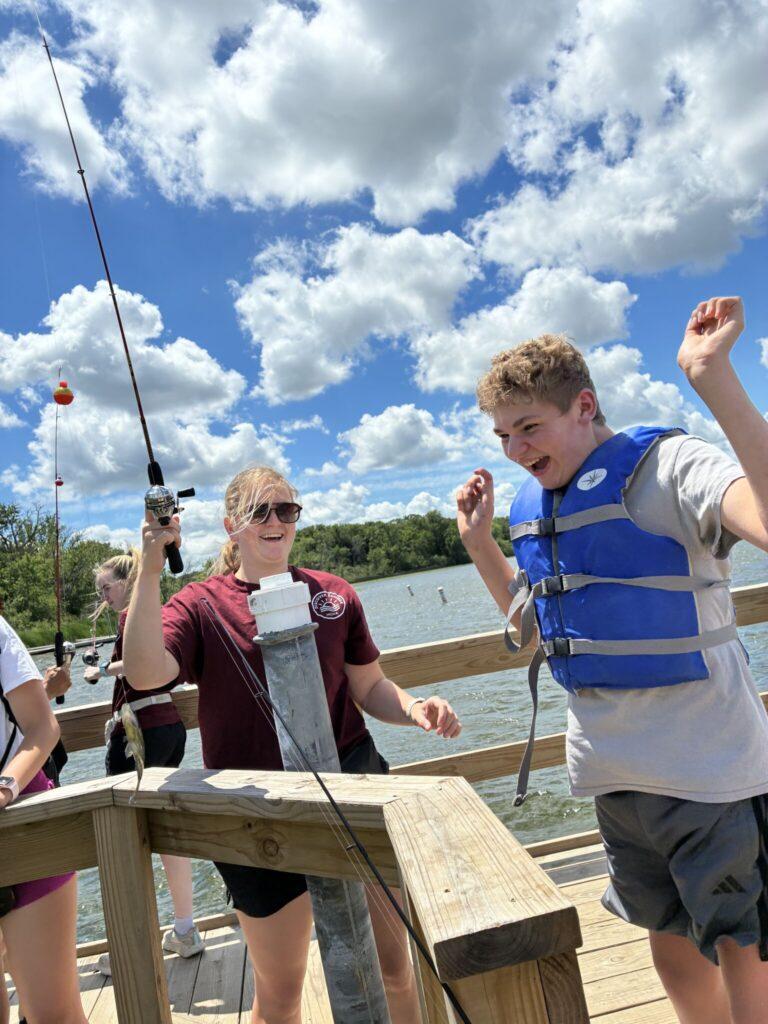 Two people wearing life jackets stand on a wooden dock by the water, one with arms raised in excitement as a fishing rod with an orange bobber lies nearby.