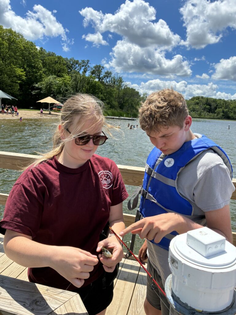Two children on a wooden dock by a lake wearing life jackets, examining a rope near a white container.