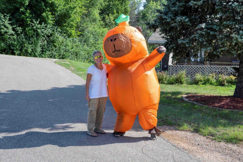A person wearing an oversized orange pumpkin mascot costume stands on a paved road next to another person, with trees in the background.