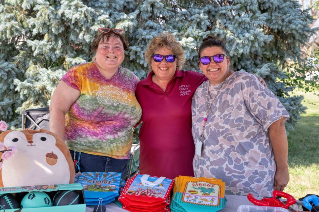 Three women stand outdoors behind a table of colorful cakes, with a panda decoration on the left.