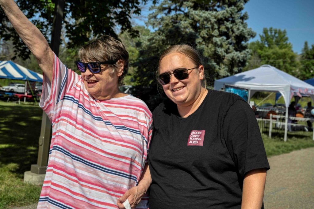 Two smiling women wearing sunglasses stand outdoors at a sunny event, one in a striped pink shirt with her arm raised and the other in a black T-shirt.