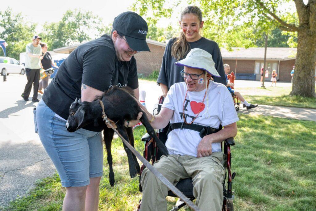 A man in a wheelchair sits with a black service dog as two people smile nearby on a sunny day.