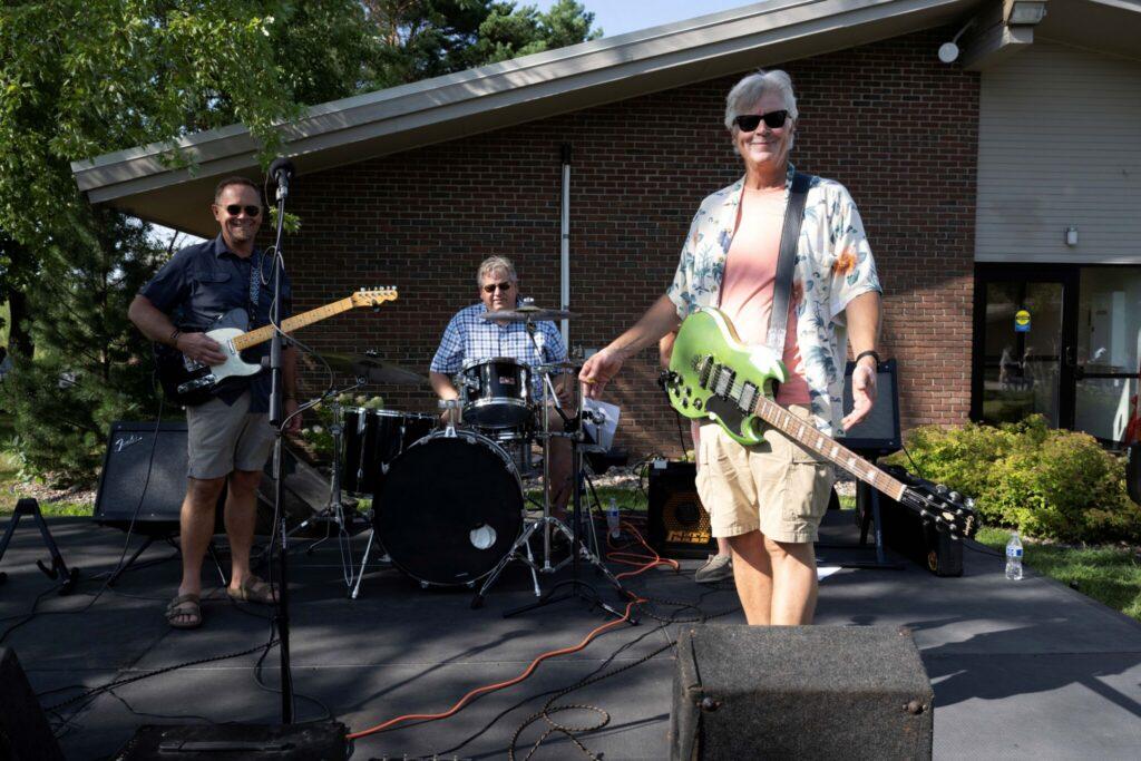 A three-member band performing outdoors on a small stage in front of a brick building, with two guitarists (one playing a bright green guitar) and a drummer.