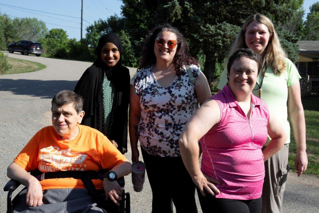 Five people posing outdoors on a sunny day, including a person in an orange shirt in a wheelchair on the left and four standing, all smiling.