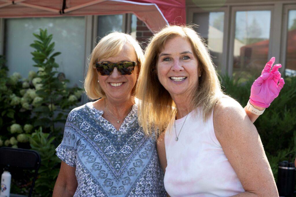 Two smiling women posing outdoors, one in a blue patterned blouse with sunglasses and the other in a light pink sleeveless top.