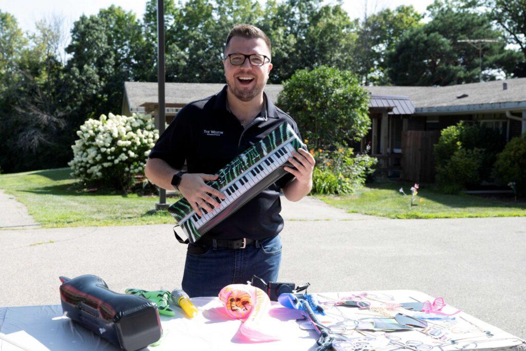 A smiling man in a dark polo shirt stands in a sunny suburban driveway holding a small keyboard.