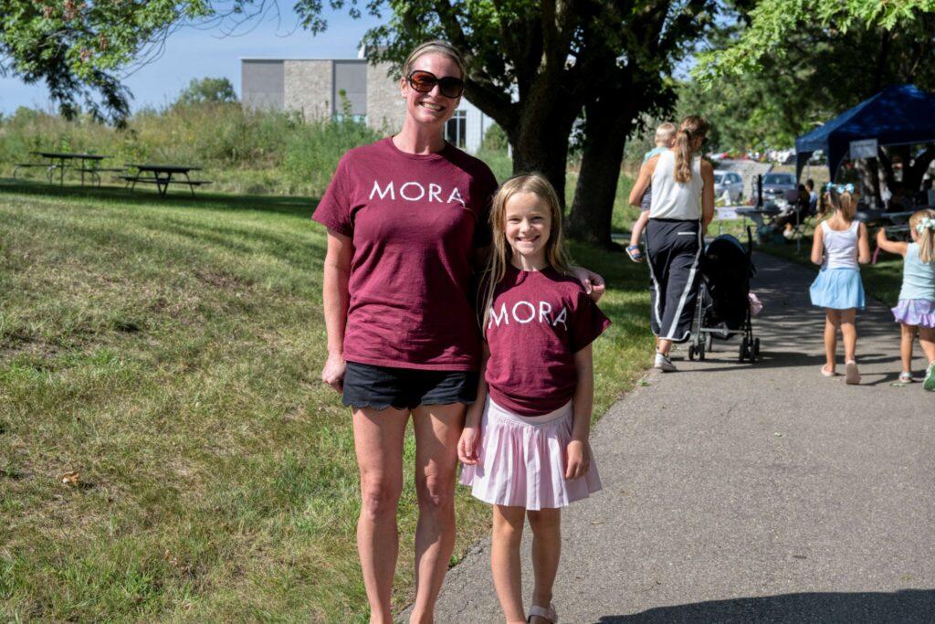 A woman and a girl wearing matching maroon shirts walk together along a sunny park path, with people and a stroller in the background.