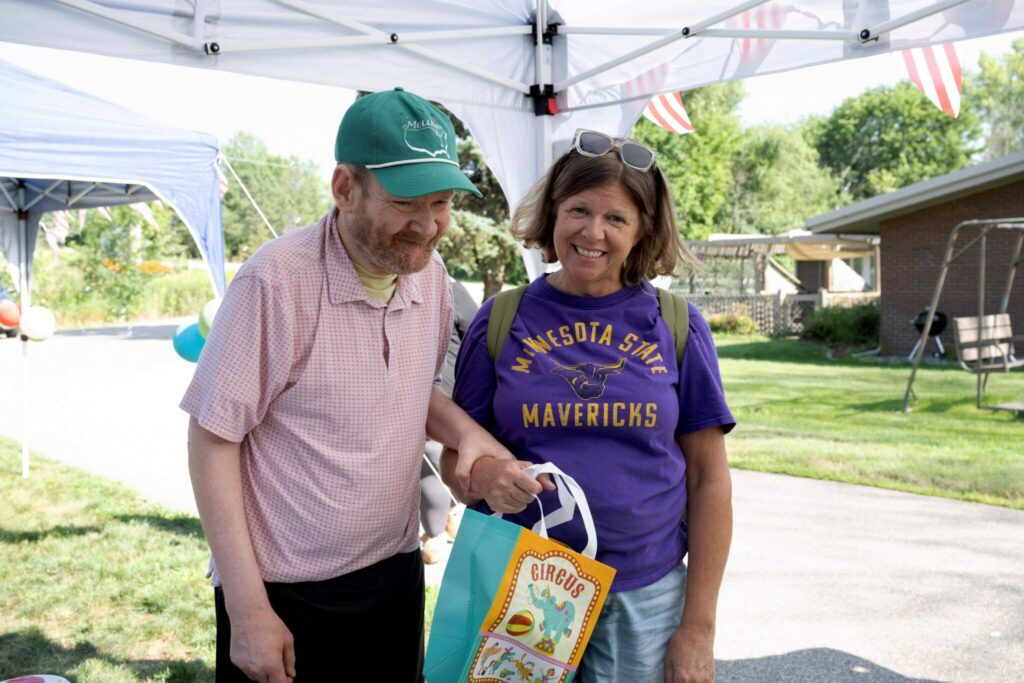 A man in a green cap and pink polo and a woman in a purple Mavericks shirt smile while looking at a colorful gift bag under a canopy.