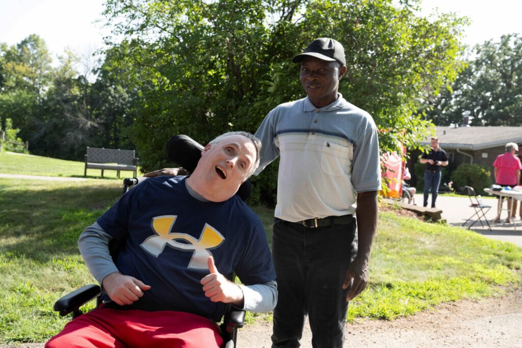 A person in a wheelchair gives a thumbs-up beside a standing man in a sunny park.