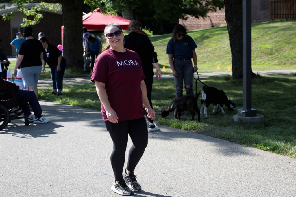 A person wearing a maroon shirt and sunglasses walks along a paved park path, with other people and dogs visible in the background.