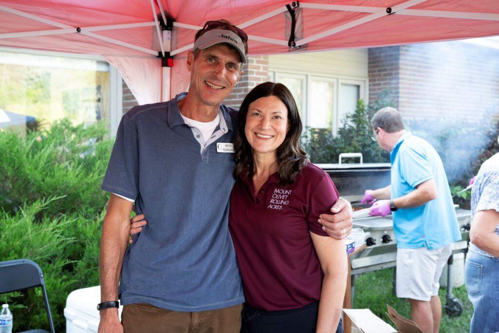 Smiling man and woman stand arm-in-arm under a red canopy at an outdoor barbecue.