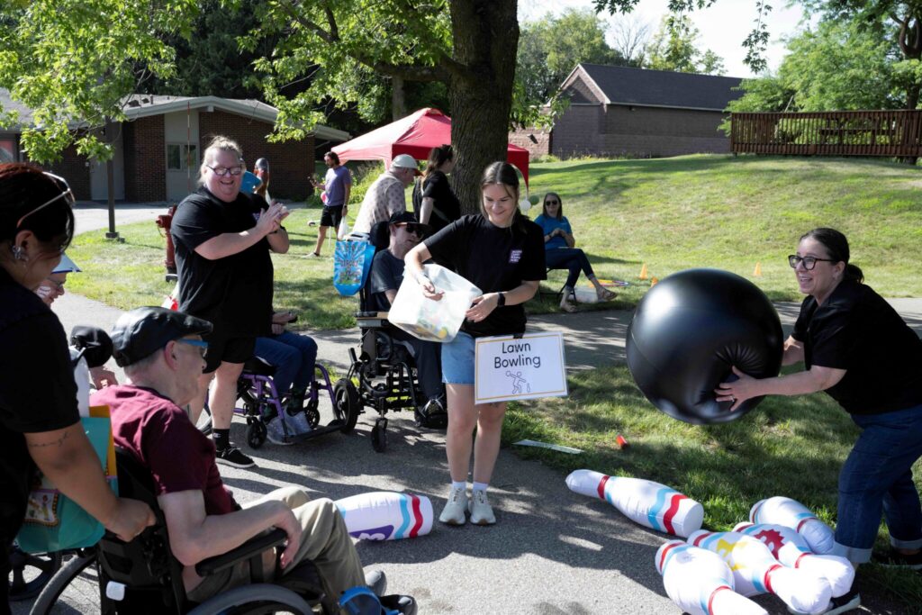 People gathered outdoors near a tree in a park, including a person in a wheelchair and someone holding a sign, with a large black exercise ball and striped foam rollers laid on the ground.