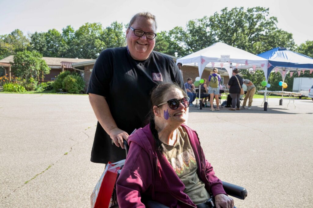 A smiling man stands behind a woman in a wheelchair at an outdoor event with tents in the background.