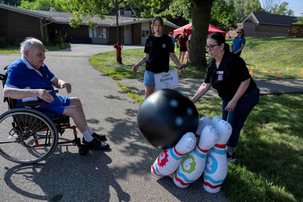 An elderly man in a wheelchair watches a game of giant outdoor bowling with a large black ball and oversized pins.