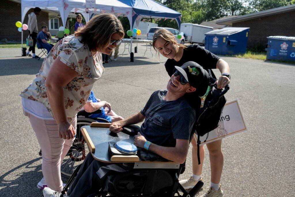 Smiling man in a wheelchair sits at a table while two women lean in to chat at an outdoor event with tents.