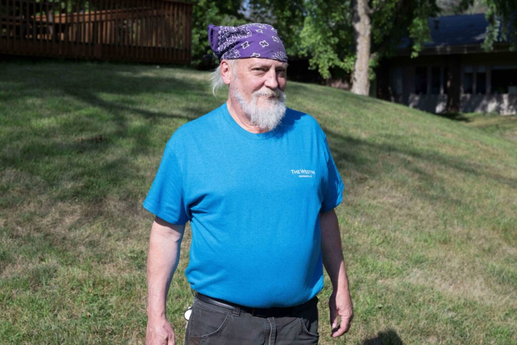 Older man with a white beard and purple bandana wearing a blue T-shirt standing on a grassy hill.