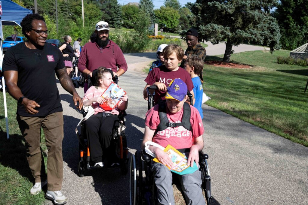 A diverse group of people walking together on a sunny park path, with some in wheelchairs.