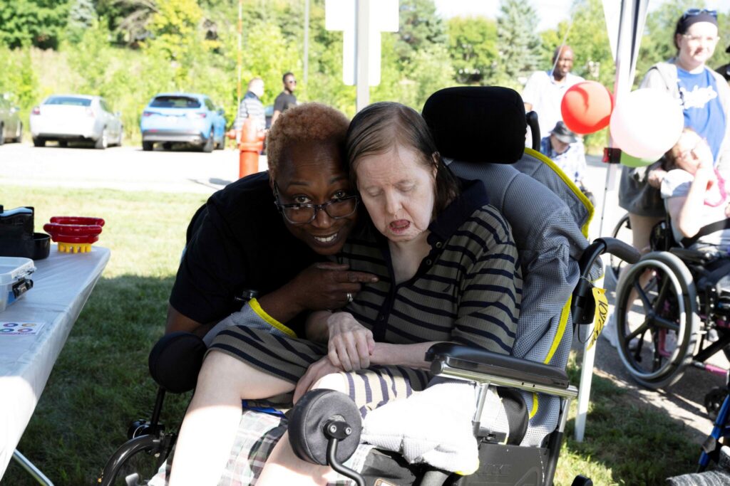 An elderly woman in a wheelchair sits with a man leaning over from behind, both focused on a small device on a table at an outdoor event.