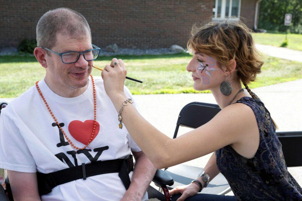 A man wearing a white I heart NY shirt sits on a park bench as a woman applies face paint to his cheek.