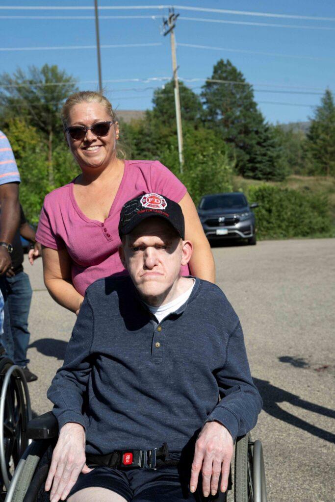 A man in a wheelchair wearing a dark blue sweater and cap sits outdoors while a smiling woman in a pink shirt stands behind him.