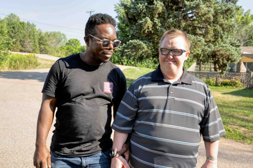 Two men wearing sunglasses stand together on a sunny suburban street with trees in the background.