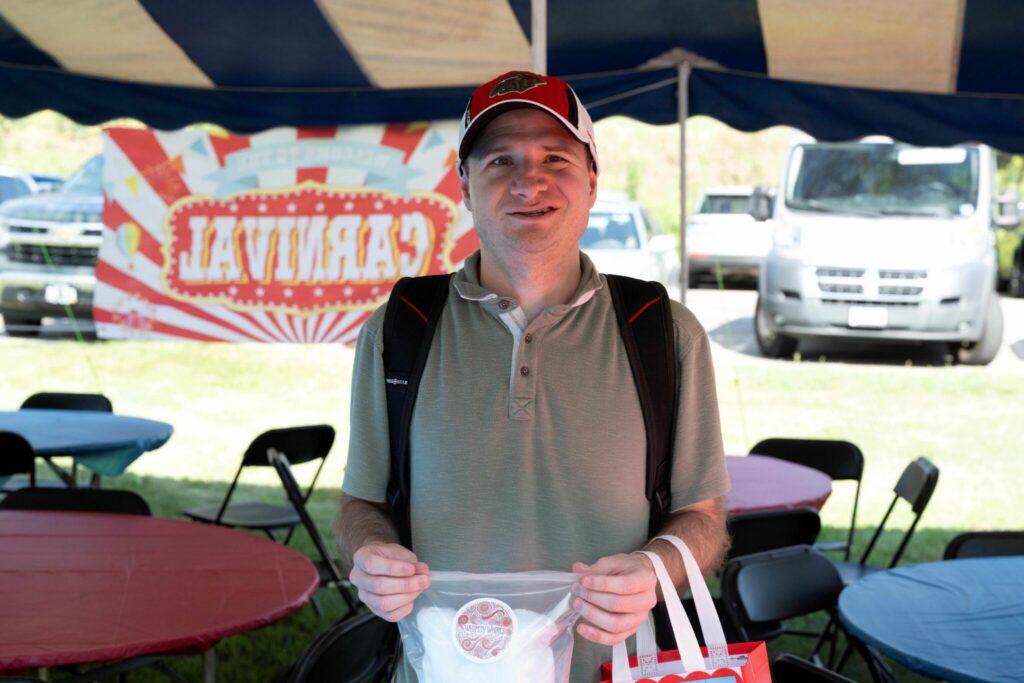 Smiling man in a khaki polo shirt and red cap stands under a blue-and-white striped tent at an outdoor event, holding a white plastic bag.