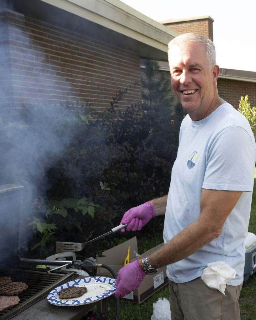 Smiling man in a light blue shirt wearing pink gloves grills burgers at an outdoor barbecue in a backyard.