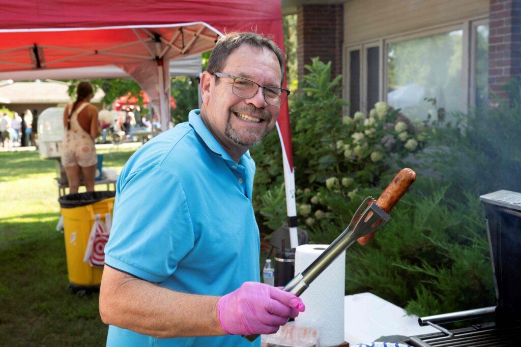 A man in a blue polo shirt wearing a pink glove grills food with tongs at an outdoor event under a red canopy, smiling at the camera.