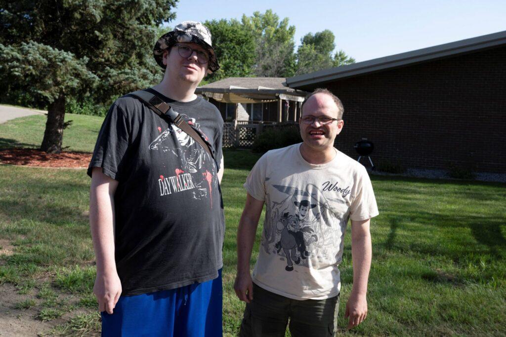 Two men stand side by side on a grassy lawn with trees and a dark building in the background.
