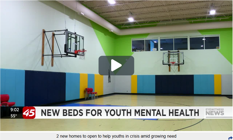 Indoor gym with padded blue, yellow, and green walls and two basketball hoops, with a video play button overlay.