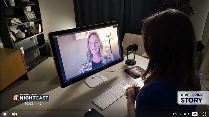 A person sits at a desk with a keyboard and mouse, watching a video call on a computer monitor that shows a woman speaking.